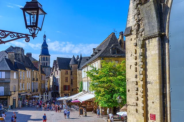Die Place de la Liberté in Sarlat-la-Canéda mit honigfarbenen Steinhäusern, einem Glockenturm in der Ferne und einer schmiedeeisernen Laterne unter blauem Himmel